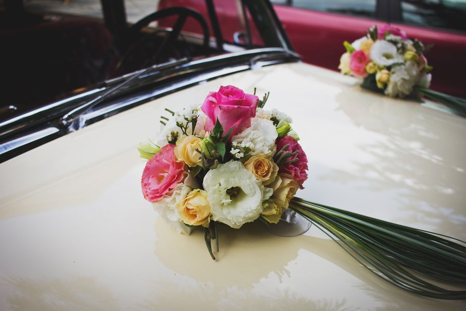 A floral bouquet featuring pink roses, white lisianthus, yellow spray roses, and pink eustoma, arranged with sprigs of greenery and wrapping paper. The bouquet is positioned on the hood of a vintage w