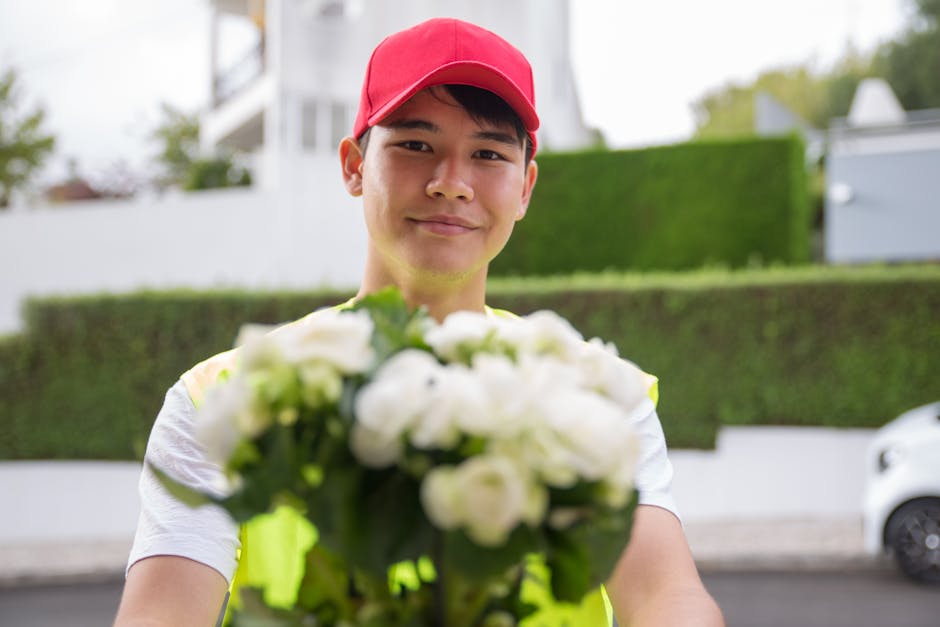 A delivery person wearing a red cap and a high-visibility yellow vest holds out a bouquet of fresh white roses and greenery towards the camera. The flowers are arranged in a compact, rounded floral gi