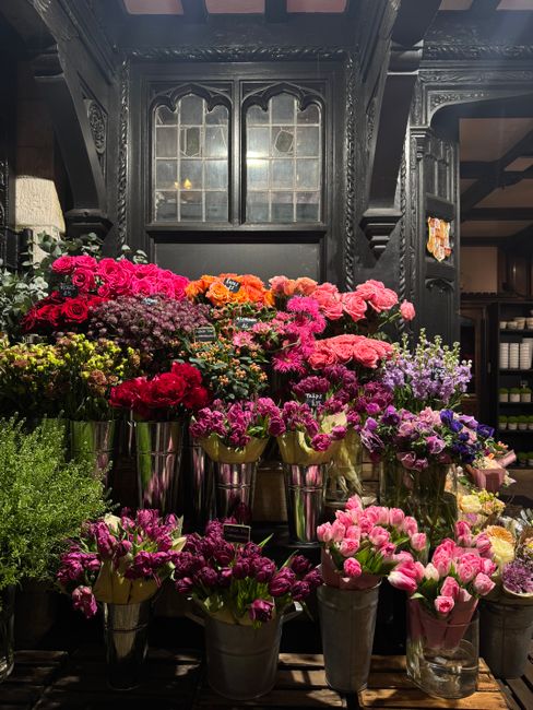 An indoor floral display featuring an array of fresh flowers including pink, purple, red, and orange blooms, arranged in silver and glass vases on a dark wooden surface. The flowers, such as hydrangea