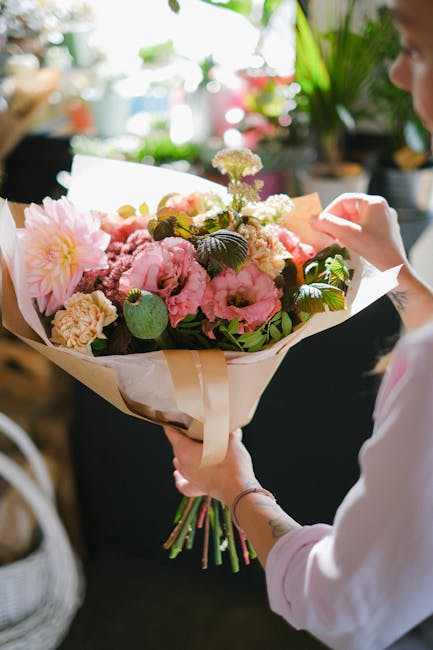 A person holding a large bouquet of fresh flowers, including pink peonies, white carnations, and small purple blossoms, wrapped in beige and white tissue paper with a satin ribbon, against a softly bl