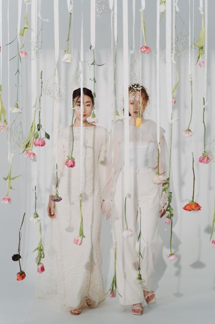 Two women are standing behind a vertical installation of suspended pink and red roses with green stems and leaves, arranged in a cascading manner against a white backdrop with sheer curtains. They are